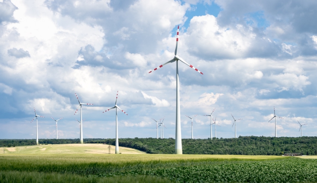 640x369-oekostrom-kraftwerk Windräder stehen majestätisch auf einer riesigen grünen Wiese unter einem wolkigen Himmel und speisen effizient saubere Energie in das Stromnetz ein.