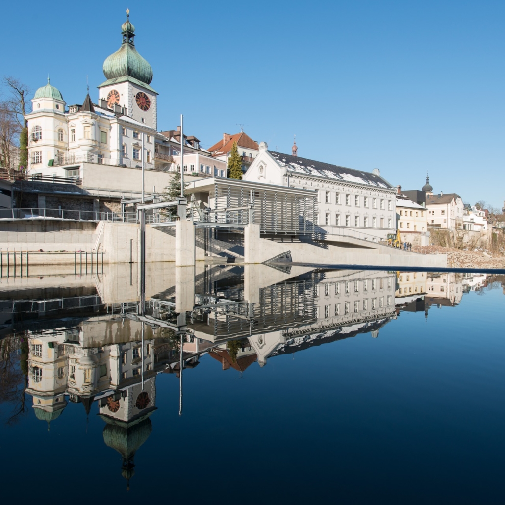 Wasserkraftwerk Waidhofen an der Ybbs, Foto: oekostrom AG Ein heiterer Blick auf die Uferpromenade einer Stadt mit historischen Gebäuden und einem Uhrenturm, der sich im ruhigen Wasser unter einem klaren blauen Himmel spiegelt, wo das Flüstern der Wasserkraft mit der Ruhe der Natur harmoniert.
