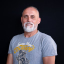 Ein Mann mit grauem Bart und kurzen Haaren trägt ein graues T-Shirt mit grafischem Muster und steht vor einem schwarzen Hintergrund.