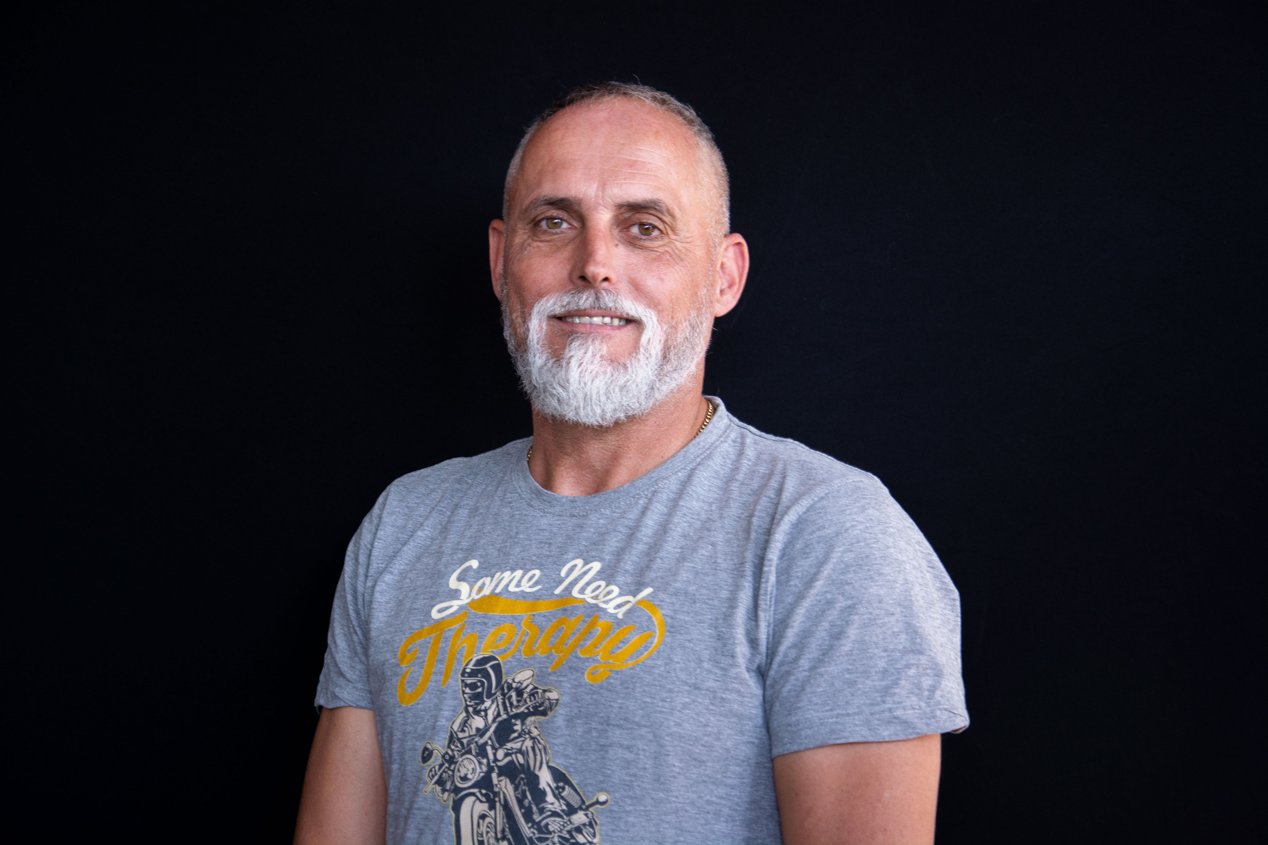 Ein Mann mit grauem Bart und kurzen Haaren trägt ein graues T-Shirt mit grafischem Muster und steht vor einem schwarzen Hintergrund.