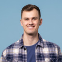 Junger Mann mit kurzen braunen Haaren, der ein blau kariertes Hemd und ein blaues T-Shirt trägt und vor einem schlichten hellblauen Hintergrund lächelt.