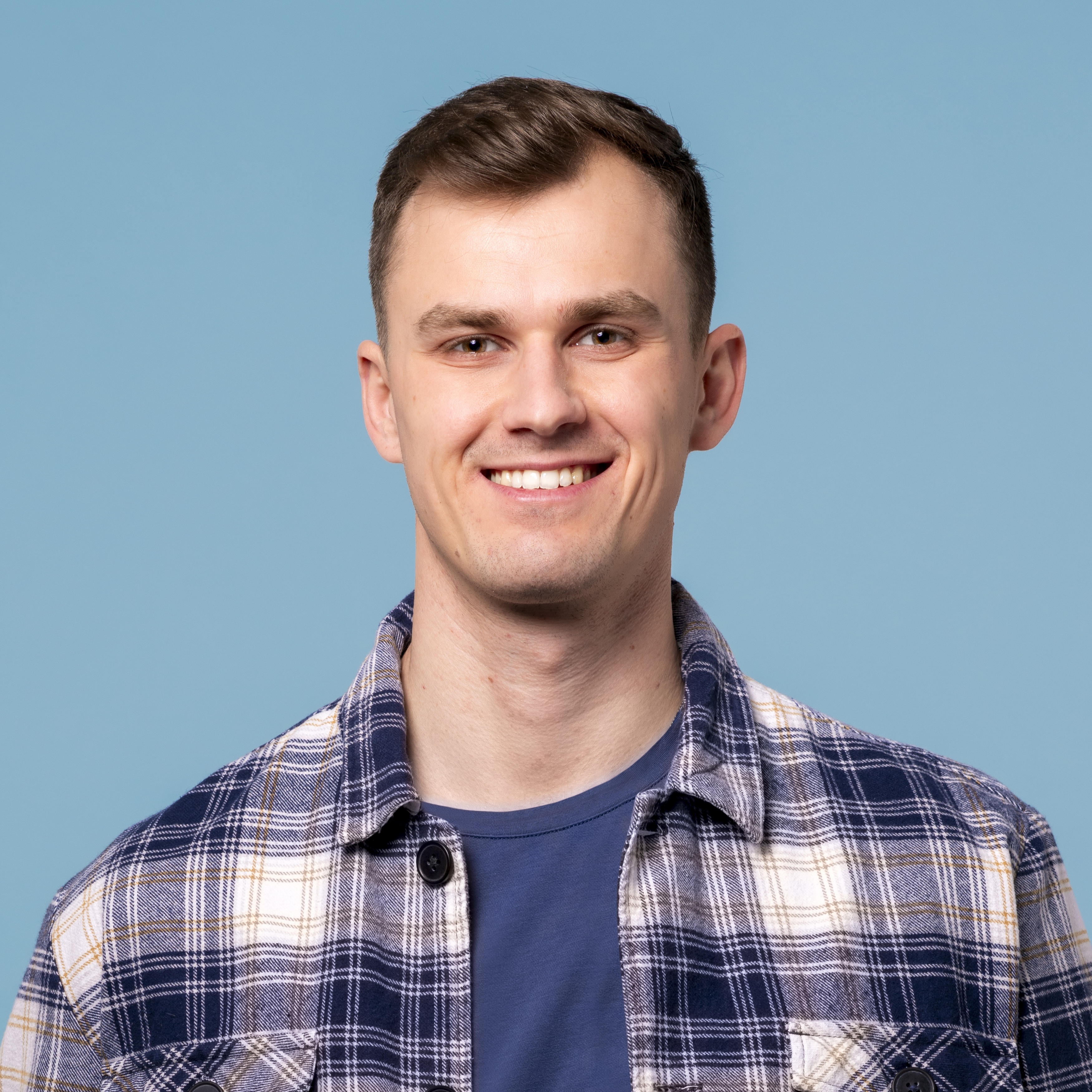 Junger Mann mit kurzen braunen Haaren, der ein blau kariertes Hemd und ein blaues T-Shirt trägt und vor einem schlichten hellblauen Hintergrund lächelt.