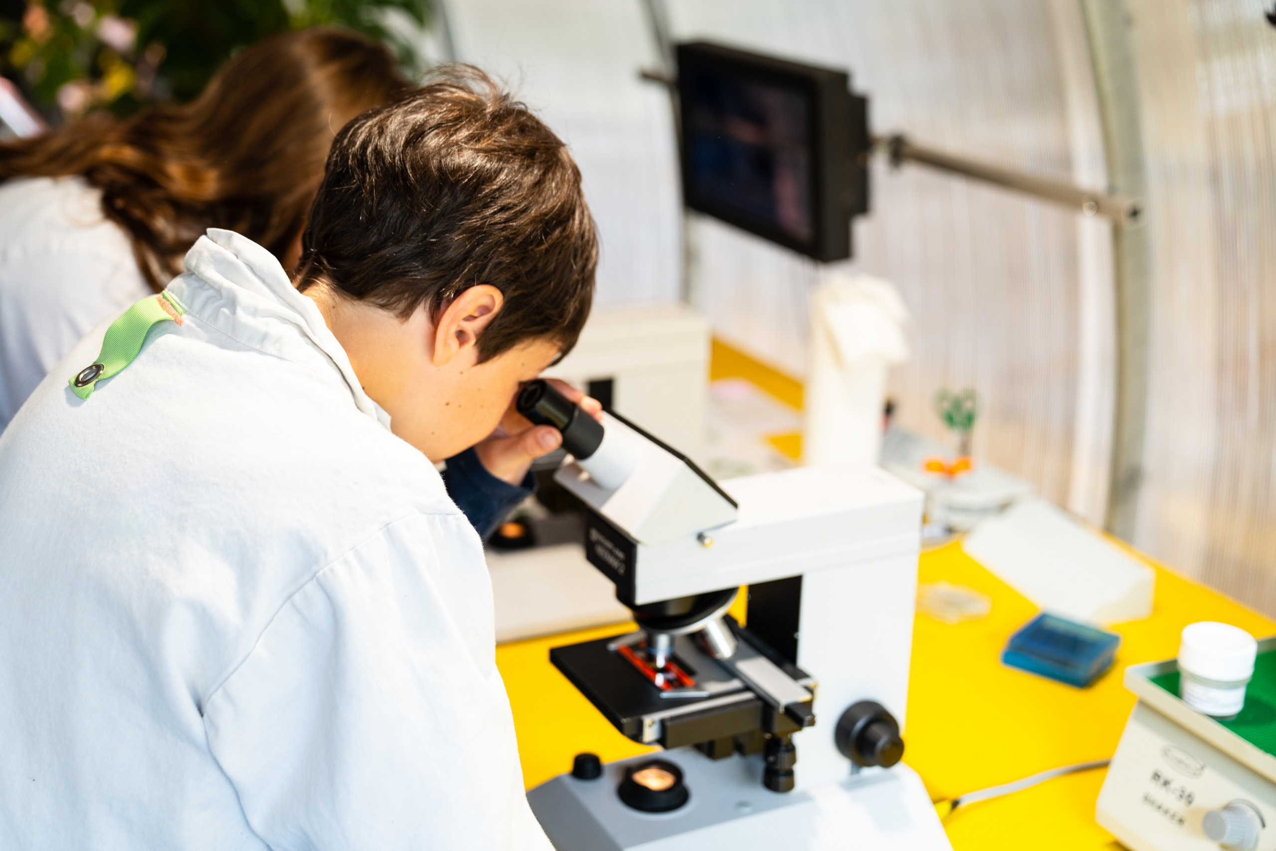 Kinder tragen einen weißen Forscherkittel und sehen durch ein Mikroskop im ZOOM Kindermuseum