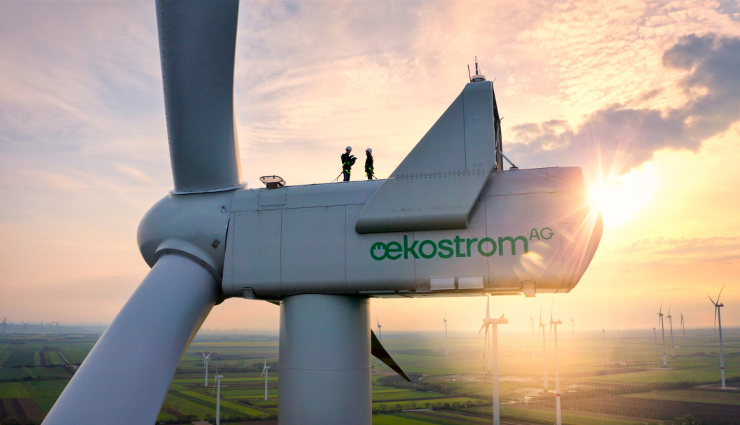 oekostrom AG: Zwei Arbeiter stehen bei Sonnenuntergang auf einer großen Windturbine mit der Marke „oekostrom AG“ und blicken im Hintergrund auf einen Windpark mit mehreren Turbinen.