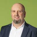Ein Mann mit Glatze und kurzem Bart, der ein weißes Hemd und einen dunklen Blazer trägt, lächelt vor einem einfarbig grünen Hintergrund.