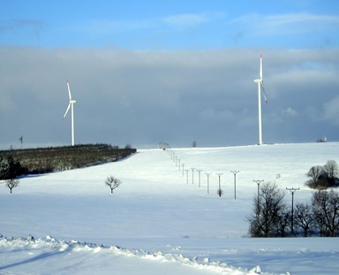 oekostrom AG: Eine verschneite Landschaft mit zwei Windrädern auf einem Hügel und einer Reihe von Strommasten, die sich in die Ferne erstrecken. Im Vordergrund sind verstreute Bäume zu sehen. Der Himmel ist teilweise bewölkt.