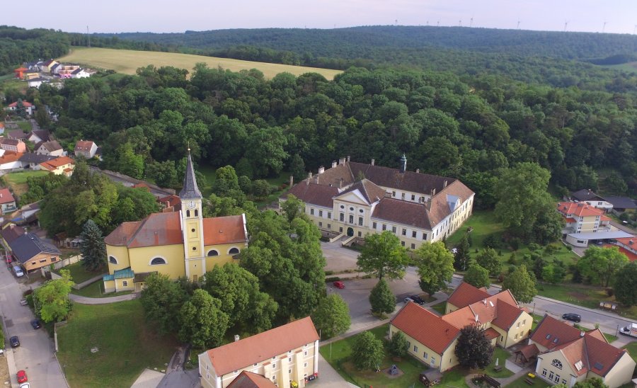 oekostrom AG: Luftaufnahme eines malerischen Dorfes mit einer gelben Kirche, einem großen Gebäude mit Innenhof und umliegenden Häusern, eingebettet zwischen dichtem Grün und sanften Hügeln.