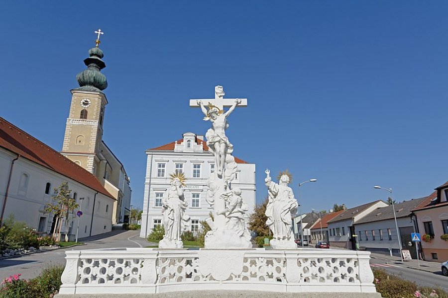 oekostrom AG: Ein Kreuzigungsdenkmal aus weißem Stein mit mehreren religiösen Statuen auf einem Stadtplatz, umgeben von Gebäuden und einer Kirche mit einer grünen Kuppel und einem Glockenturm. Im Hintergrund klarer blauer Himmel.