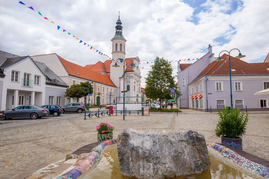 oekostrom AG: Ein Stadtplatz mit einem Brunnen mit einem steinernen Mittelstück, bunten Fahnen, geparkten Autos, Gebäuden mit roten Dächern und einer Kirche mit einem hohen Turm im Hintergrund.