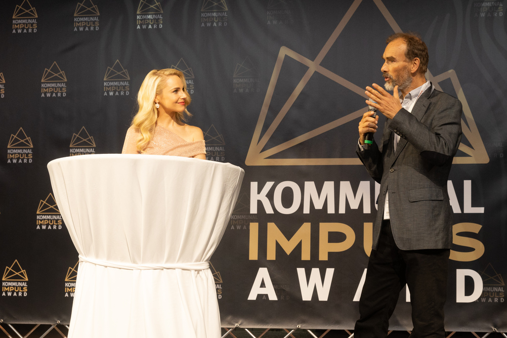 oekostrom AG: Bei der Verleihung des Kommunal Impuls Awards stehen eine Frau und ein Mann auf der Bühne. Die Frau steht hinter einem runden Tisch mit weißer Tischdecke, der Mann spricht in ein Mikrofon.