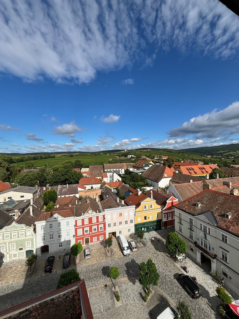 oekostrom AG: Eine erhöhte Ansicht eines malerischen Stadtplatzes mit farbenfrohen Gebäuden, Kopfsteinpflasterstraßen und ein paar geparkten Autos. Im Hintergrund sind grüne Hügel und ein blauer Himmel mit vereinzelten Wolken zu sehen.