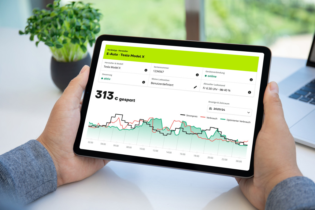 oekostrom AG: Hände halten ein Tablet, auf dem Daten eines Elektrofahrzeugs, Tesla Model X, angezeigt werden, darunter Batteriestatus und ein Diagramm des Stromverbrauchs. Im Hintergrund sind eine Topfpflanze und ein Teil eines Laptops zu sehen.