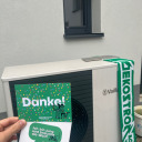 oekostrom AG: Eine Hand hält eine grüne Dankeskarte vor einer Außenanlage für Wärmepumpen neben einem Fenster. Daneben hängt ein grüner Schal mit der Aufschrift „Ökostrom“.