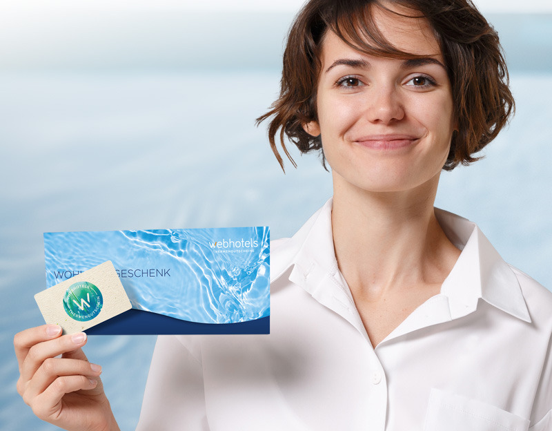 Eine Frau in einem weißen Hemd hält eine Webhotels Thermengutscheine-Geschenkkarte und einen Umschlag mit einem Wassermotiv vor einem unscharfen blauen Hintergrund.