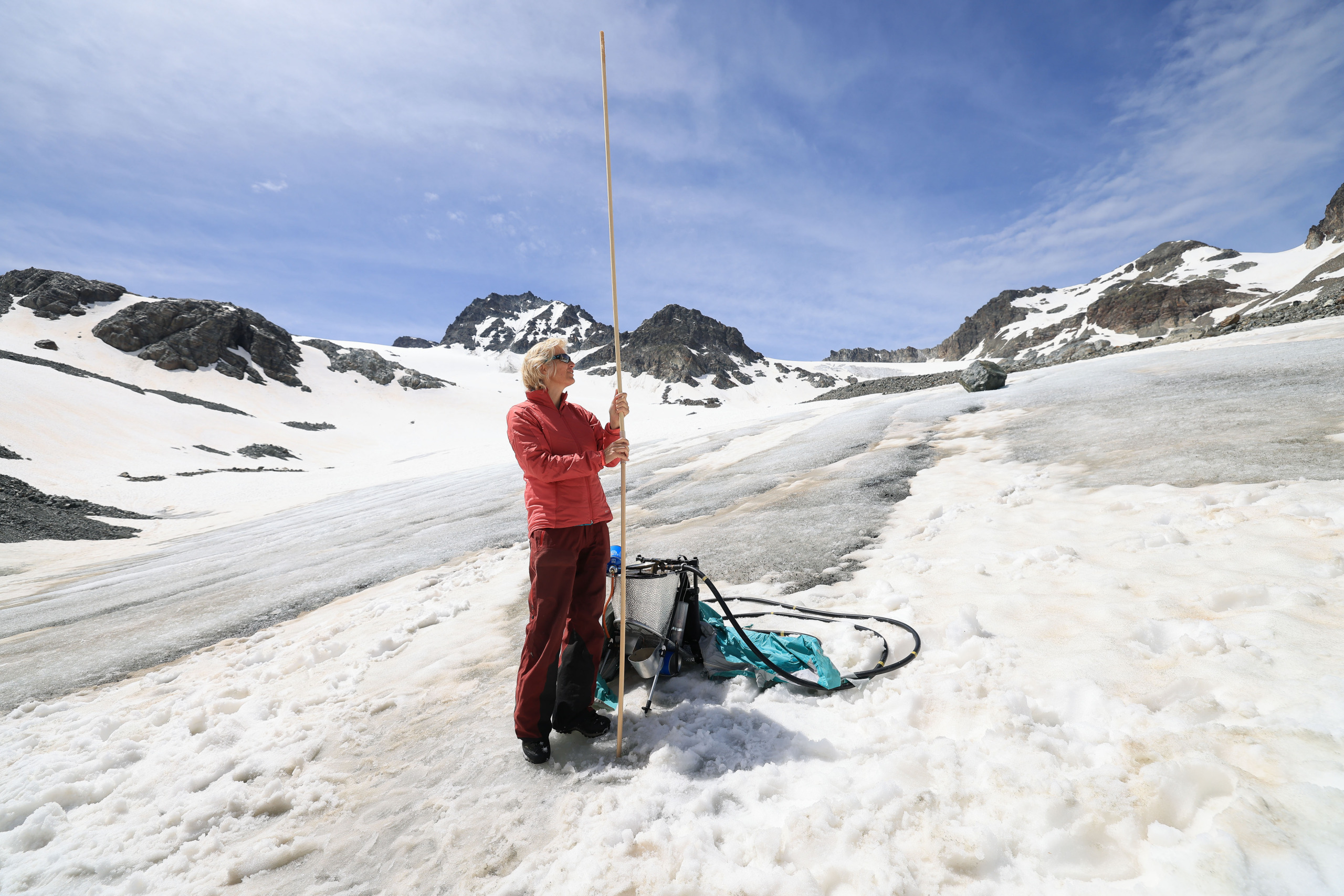 Eine Person in Rot misst mit einer langen Messlatte die Schneetiefe auf einem schneebedeckten Berggelände unter blauem Himmel und symbolisiert damit unser Engagement für den Klimaschutz. Daneben liegt die Ausrüstung, die die Bedeutung nachhaltiger Praktiken zum Schutz dieser unberührten Landschaften unterstreicht.