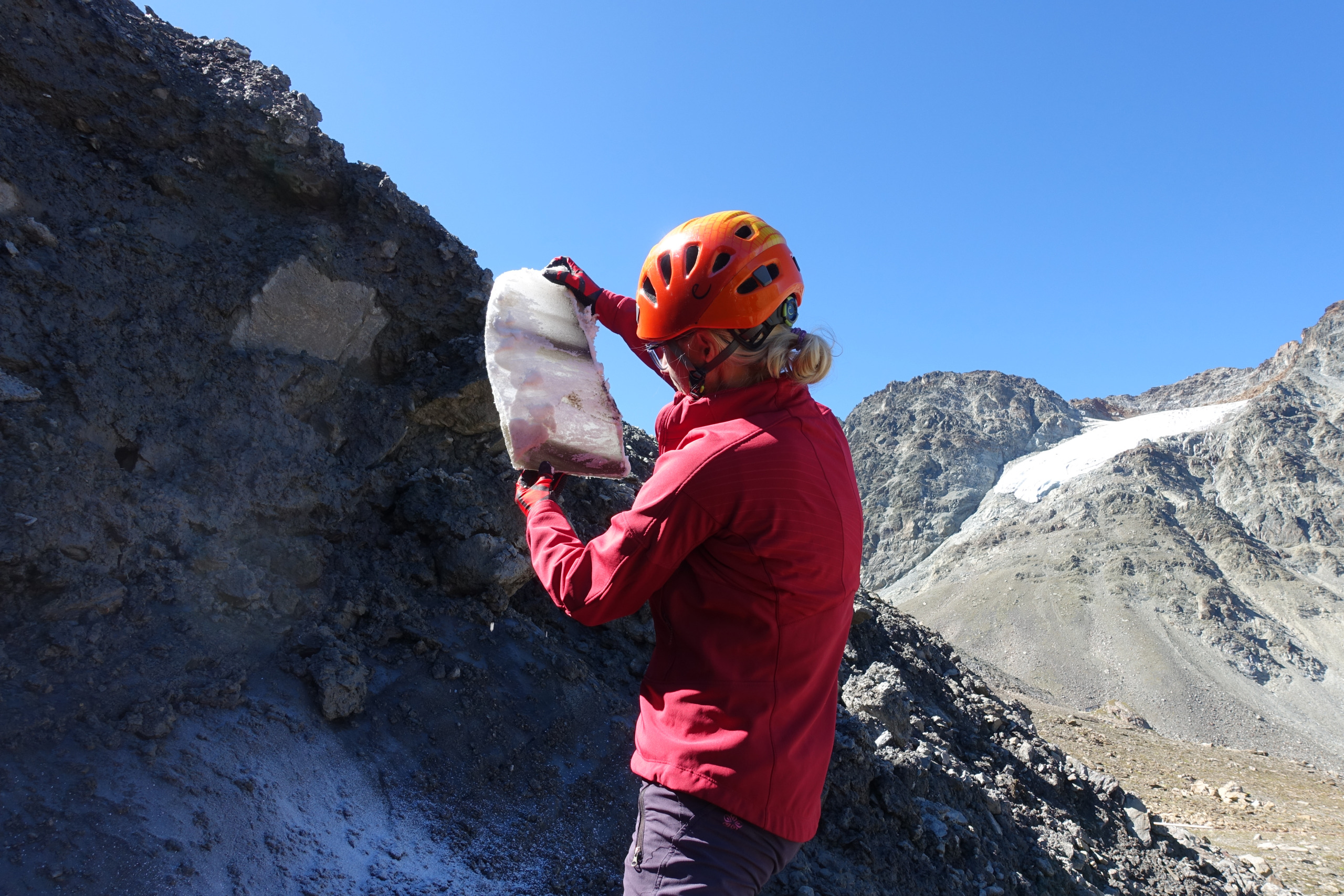 Eine Person mit rotem Helm und Jacke untersucht unter einem klaren blauen Himmel ein großes Felsstück vor einer Bergkulisse und verkörpert den Geist der Kreislaufwirtschaft, indem sie nach nachhaltigen Erkenntnissen in der Natur sucht.