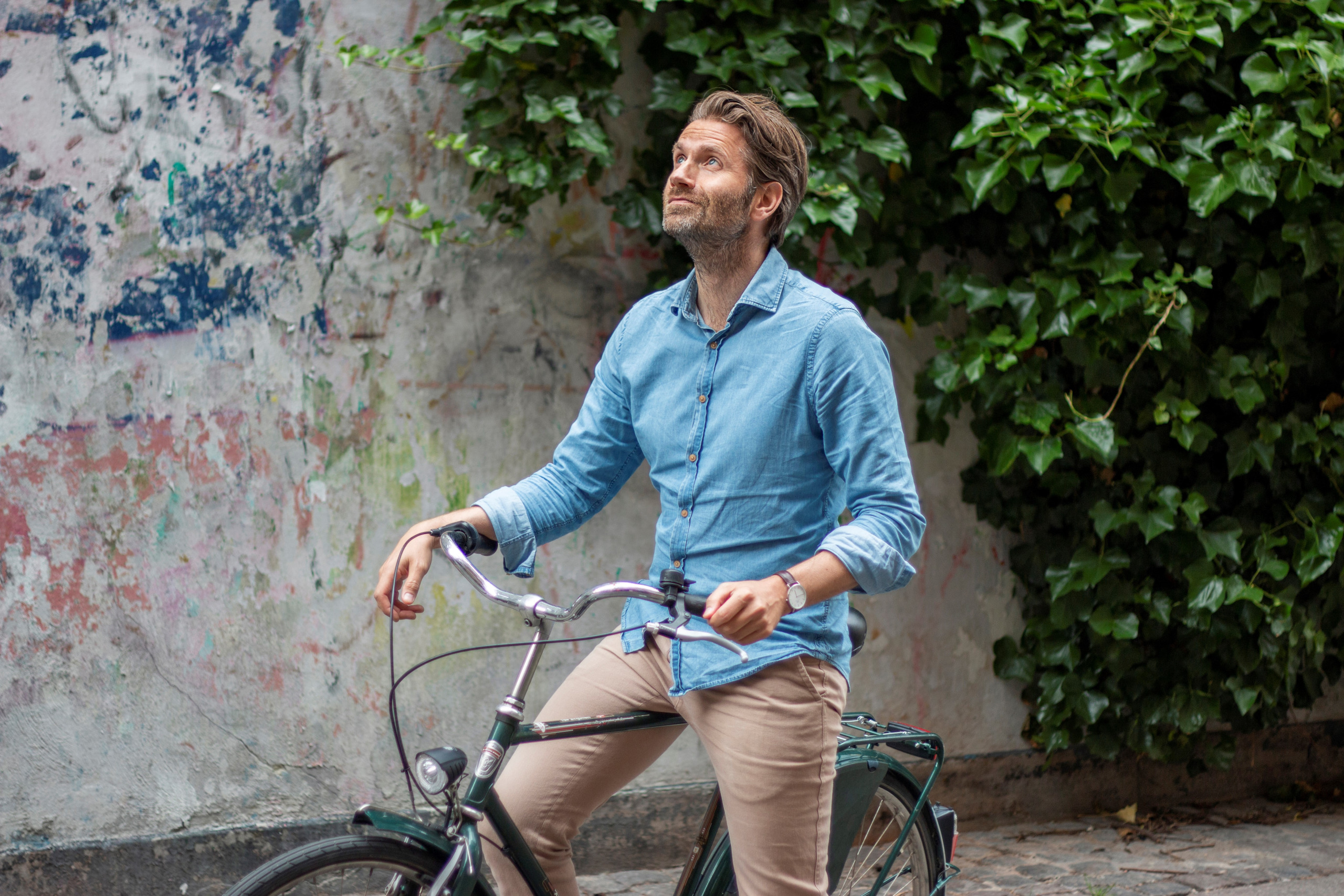 Ein Mann im blauen Hemd sitzt auf einem Fahrrad und blickt nach oben, während grünes Efeu und eine strukturierte Wand den Hintergrund bilden – eine perfekte Kulisse, um sich das Potenzial eines Balkonkraftwerks vorzustellen.