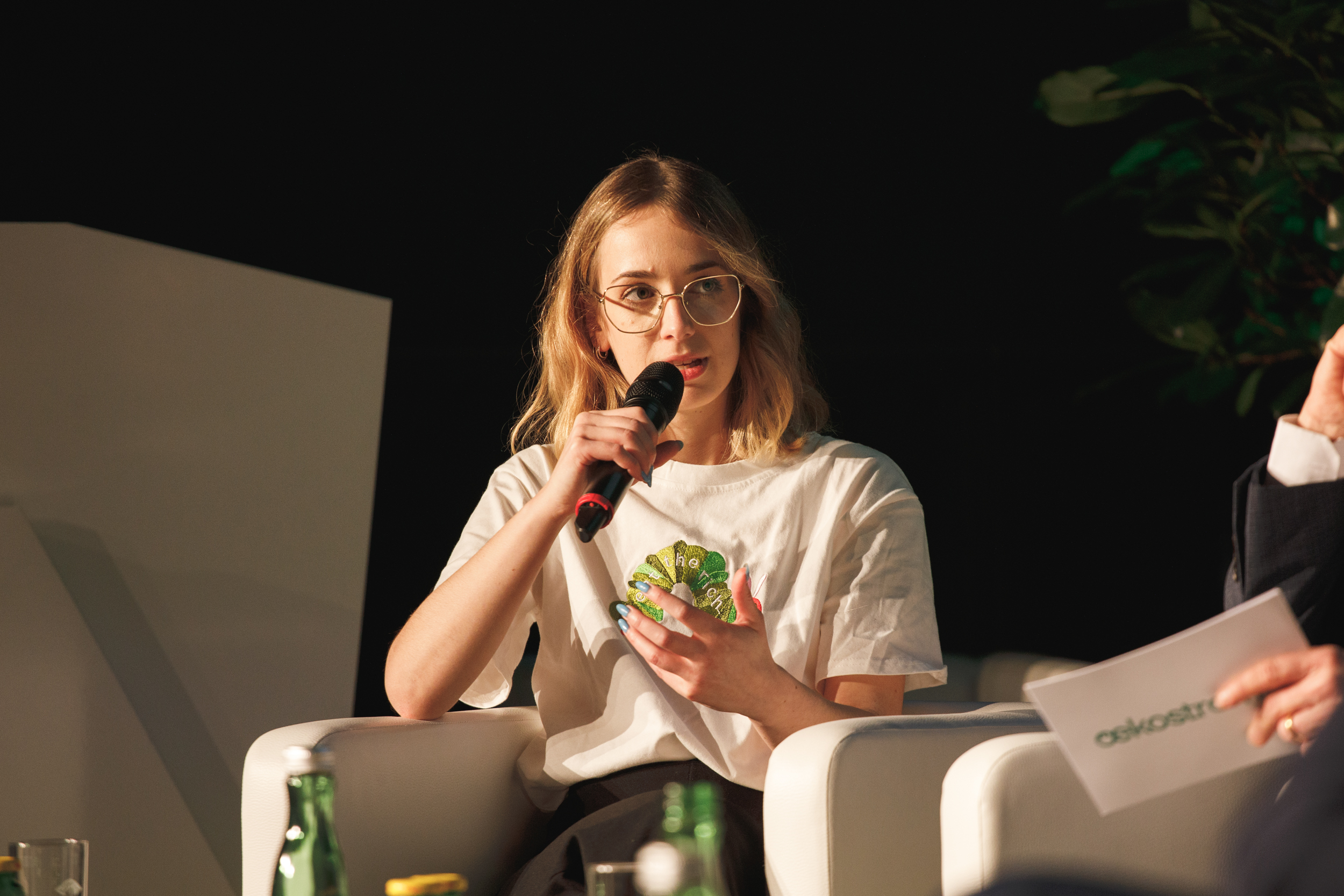 Eine Person mit Brille sitzt auf einem weißen Stuhl, trägt ein weißes Hemd und spricht in ein Mikrofon, während sie begeistert über die neuesten Innovationen auf dem Campus der Ökostrom AG diskutiert.