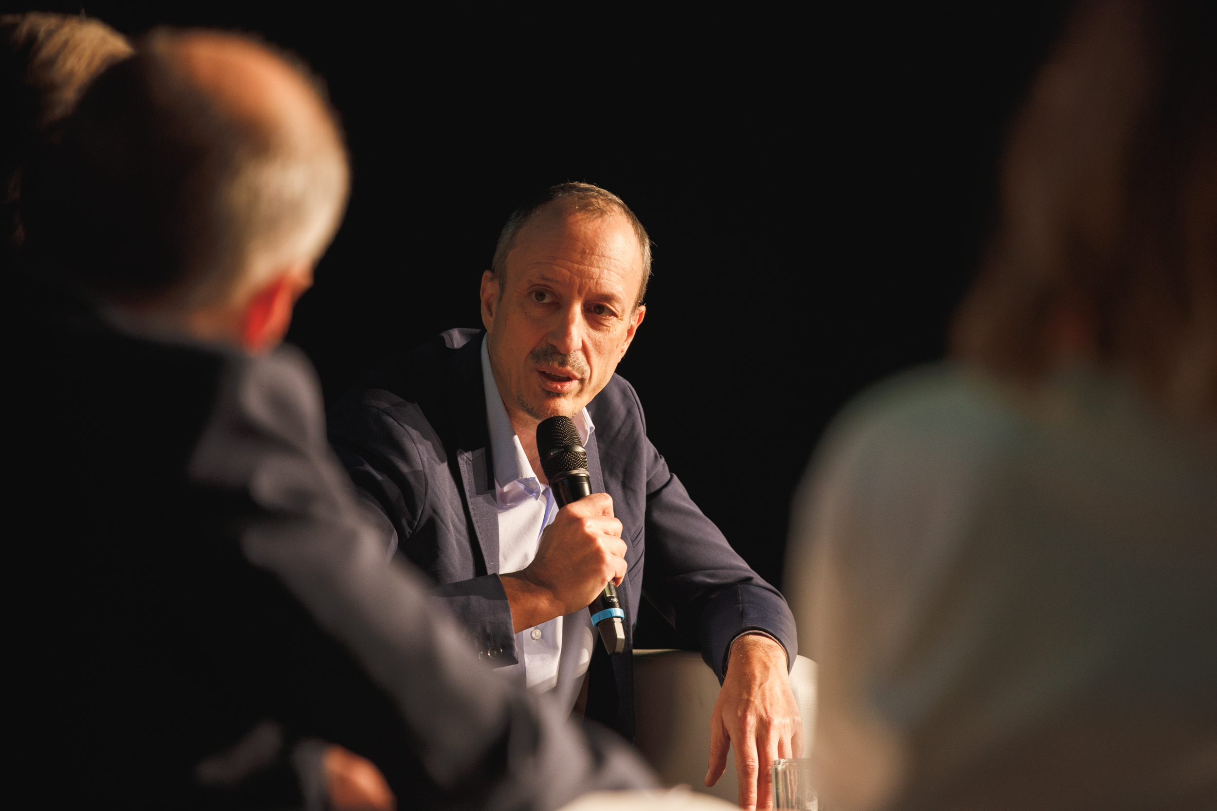 Ein Mann im Anzug spricht während einer Podiumsdiskussion zum Thema nachhaltige Energie, veranstaltet von der oekostrom AG am Campus, in ein Mikrofon. Im Vordergrund sind zwei weitere unscharfe Personen zu sehen.