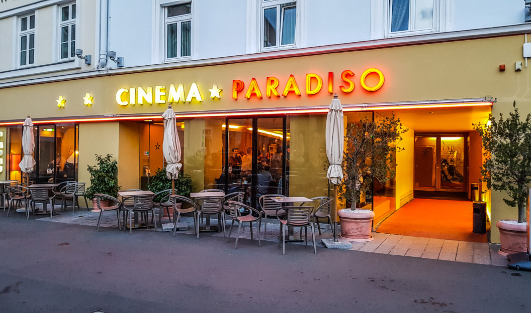 Vorderansicht des Cinema Paradiso, das jetzt mit Ökostrom betrieben wird, ein Gebäude mit gelber Fassade und Neonreklame sowie Sitzgelegenheiten im Freien mit Tischen und Stühlen unter Sonnenschirmen.