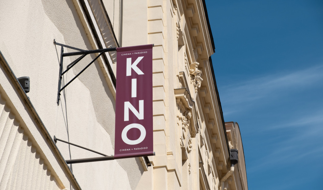 Ein vertikales „KINO“-Schild ziert die Seite eines klassischen Gebäudes und ragt vor einem Hintergrund aus leuchtendem Blau in den Himmel. Dieses Cinema Paradiso, das jetzt Ökostrom bietet, zelebriert den Charme des Kinos in einem umweltfreundlichen Stil.