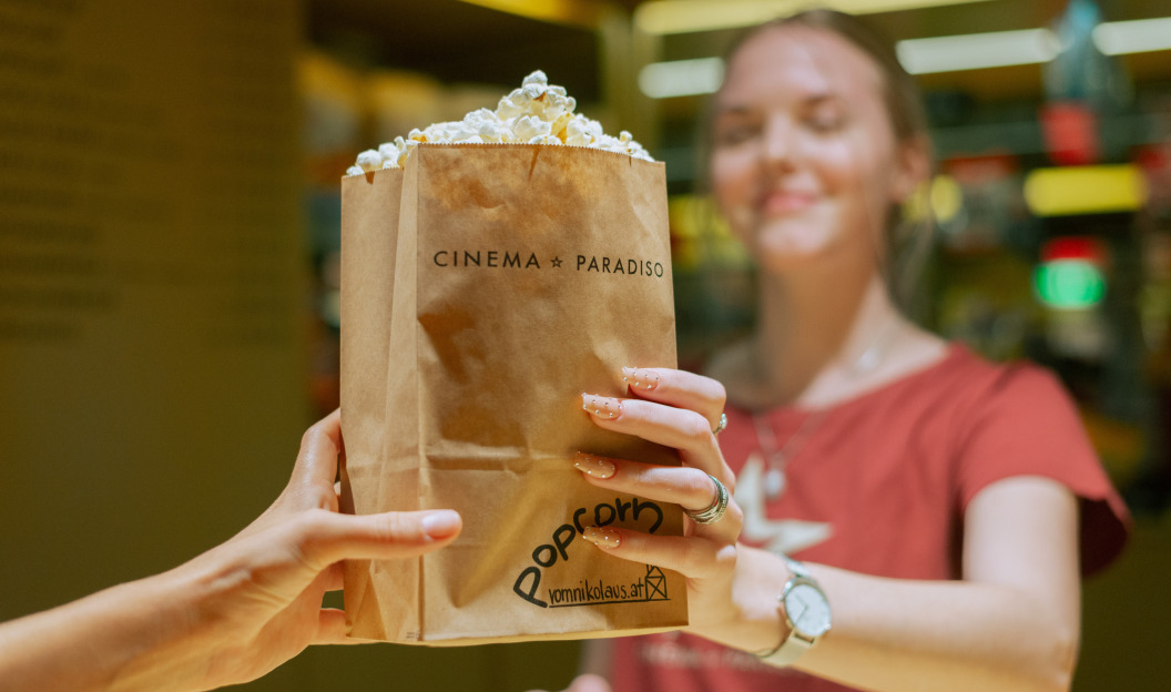 Bei einer malerischen Vorführung im Cinema Paradiso überreicht eine Person einer anderen eine braune Papiertüte mit Popcorn und genießt die elektrisierende Atmosphäre, die vollständig mit Ökostrom betrieben wird.