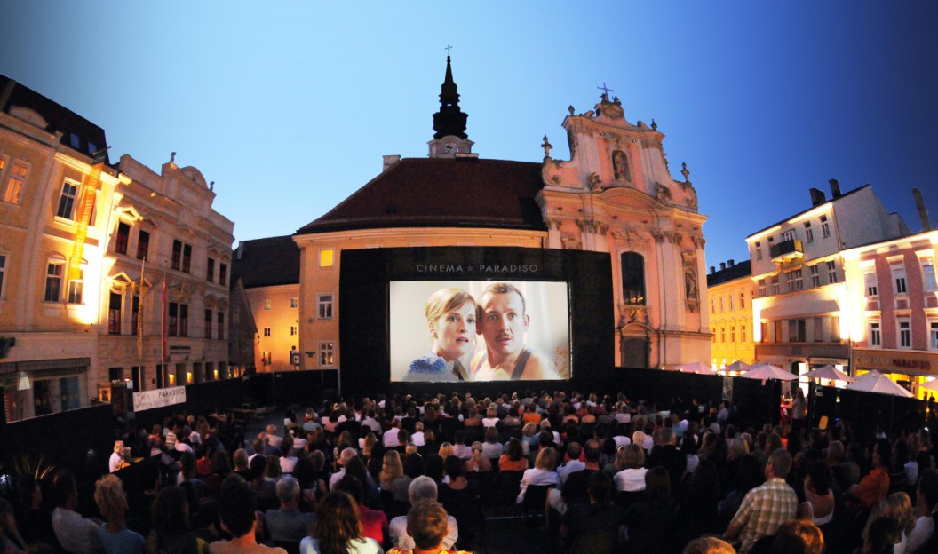 Eine große Menschenmenge versammelt sich unter klarem Himmel zu einer Freiluftvorführung von *Cinema Paradiso* auf einem europäischen Platz, umgeben von historischen Gebäuden. Die Veranstaltung findet im Zeichen des Ökostroms statt und feiert Nachhaltigkeit in dieser zauberhaften Umgebung.