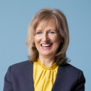 Eine lächelnde Person mit schulterlangem Haar, die eine gelbe Bluse und einen dunklen Blazer trägt, posiert vor einem schlichten blauen Hintergrund.