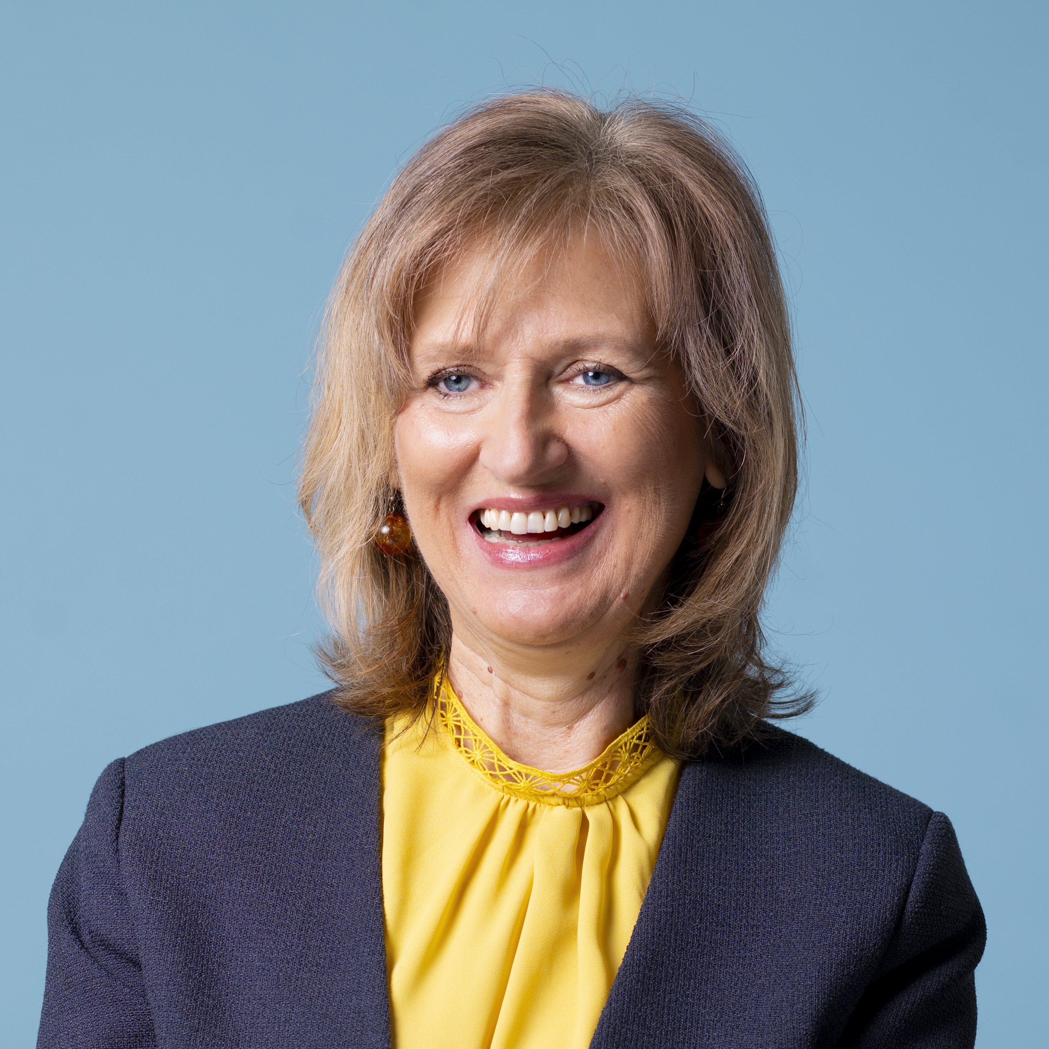 Eine lächelnde Person mit schulterlangem Haar, die eine gelbe Bluse und einen dunklen Blazer trägt, posiert vor einem schlichten blauen Hintergrund.