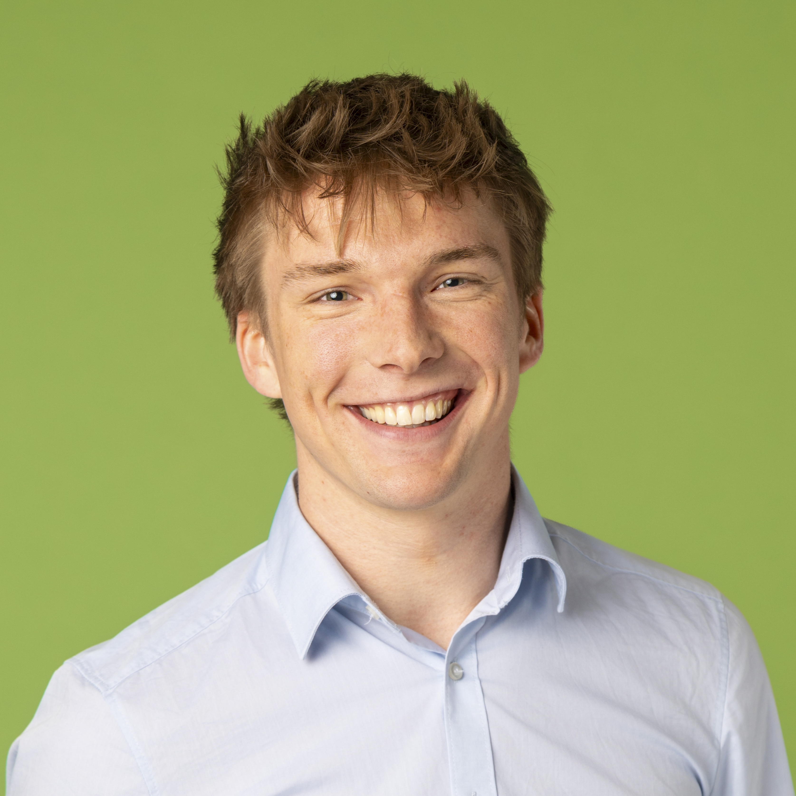 Junger Mann mit hellbraunem Haar, der ein hellblaues Button-up-Hemd trägt und vor einem schlichten grünen Hintergrund lächelt.