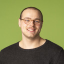 Eine Person mit kurzen Haaren, einer Brille und einem dunklen Pullover lächelt vor einem einfarbig grünen Hintergrund.