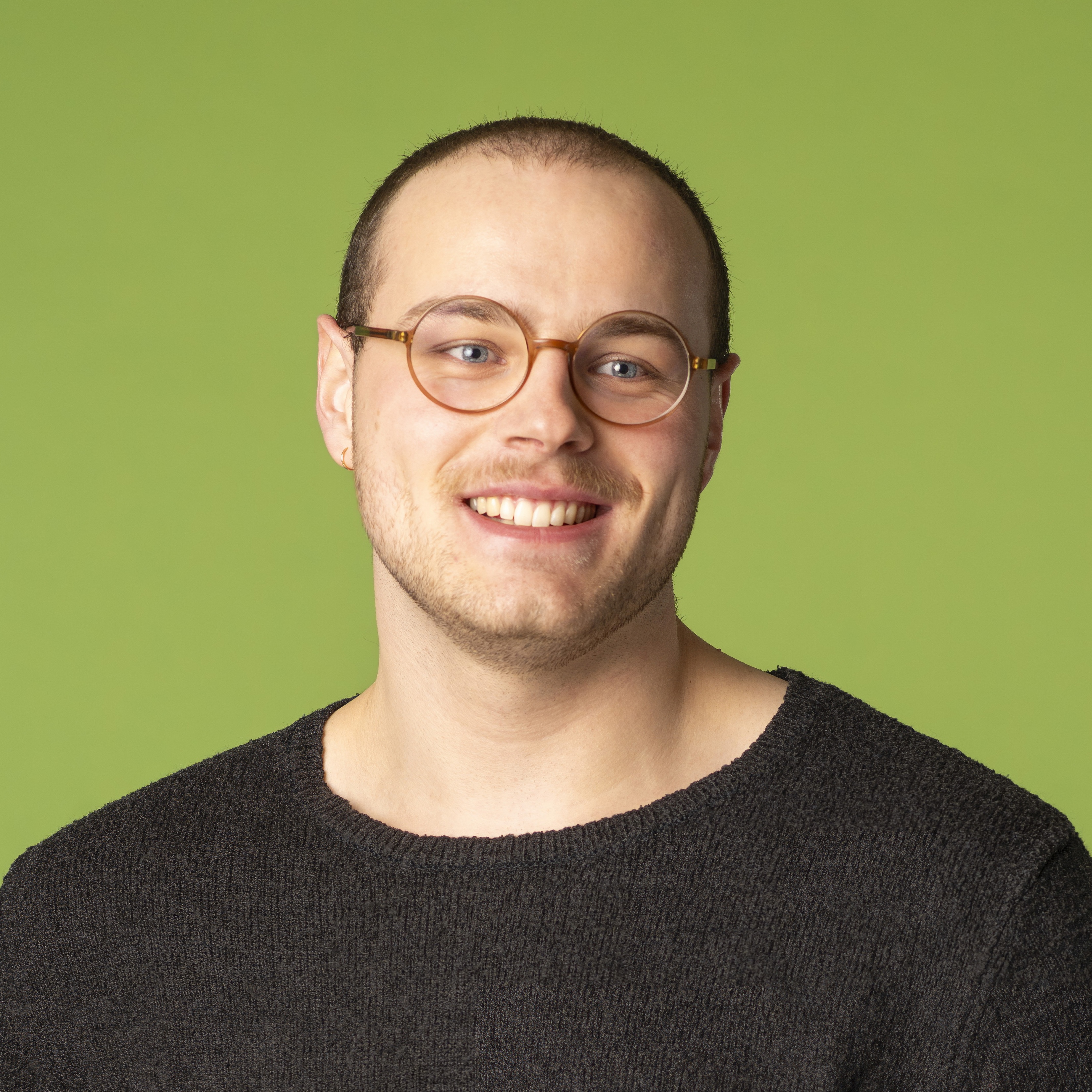 Eine Person mit kurzen Haaren, einer Brille und einem dunklen Pullover lächelt vor einem einfarbig grünen Hintergrund.