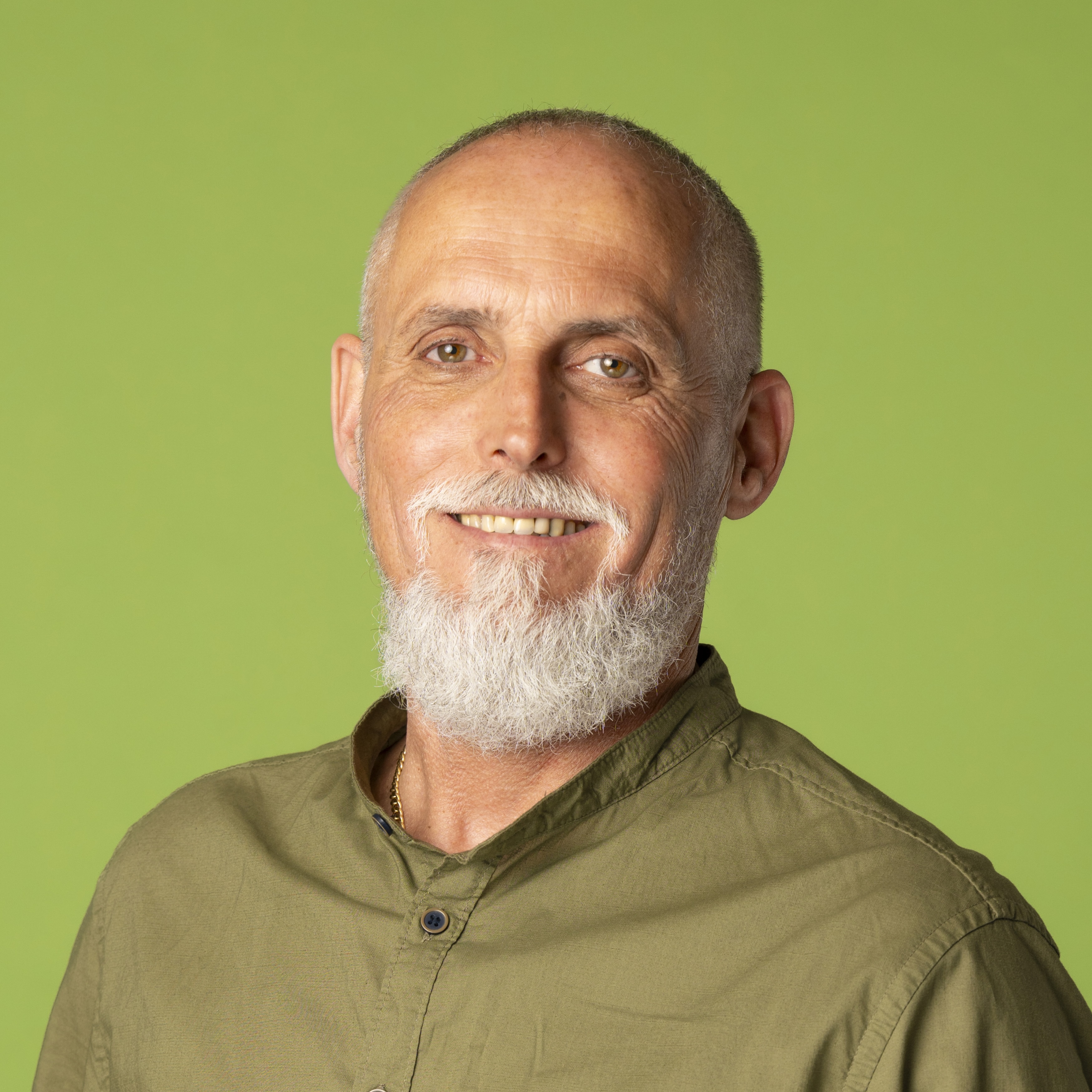 Ein Mann mittleren Alters mit kurzem grauem Bart und rasiertem Kopf, der ein grünes Hemd mit Knöpfen trägt, lächelt in die Kamera vor einem schlichten hellgrünen Hintergrund.