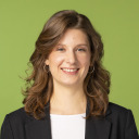 Eine Frau mit gewelltem braunem Haar, die Ohrringe, ein weißes Oberteil und einen schwarzen Blazer trägt, lächelt in die Kamera vor einem einfarbigen grünen Hintergrund.