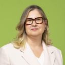 Eine Frau mit schulterlangem blondem Haar und Brille trägt einen hellen Blazer und ein weißes Oberteil und steht vor einem grünen Hintergrund.