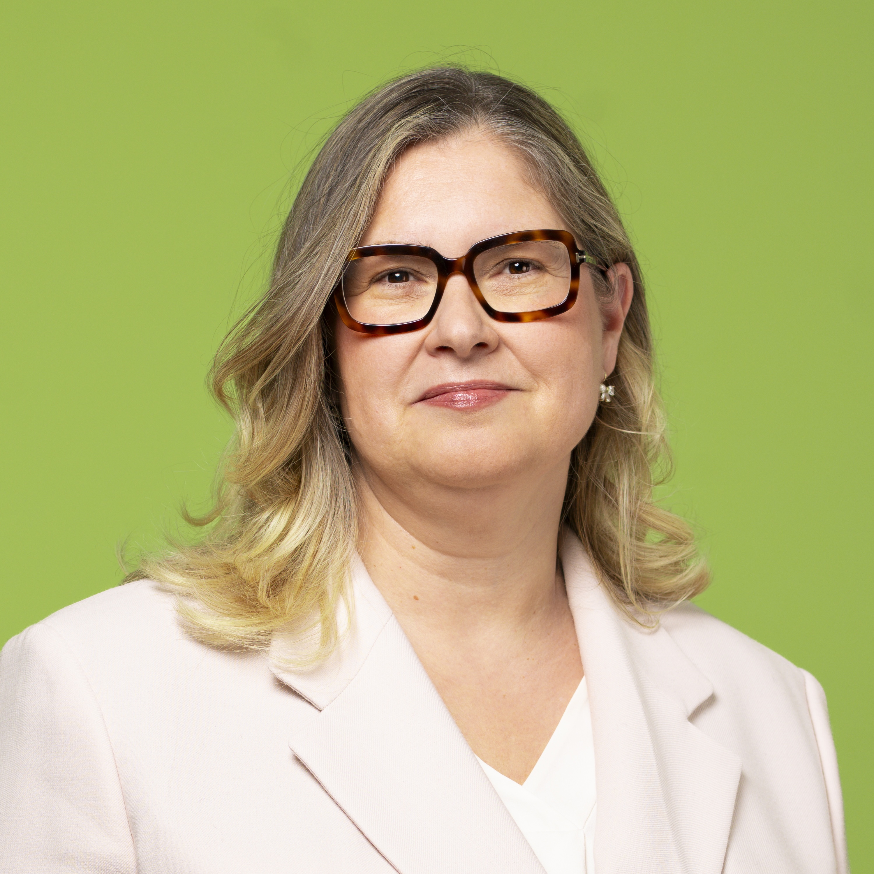Eine Frau mit schulterlangem blondem Haar und Brille trägt einen hellen Blazer und ein weißes Oberteil und steht vor einem grünen Hintergrund.