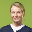 Frau mittleren Alters mit hellen, nach hinten gezogenen Haaren, die einen marineblauen Pullover über einem weißen Hemd trägt, steht vor einem einfarbig grünen Hintergrund, blickt in die Kamera und lächelt leicht.