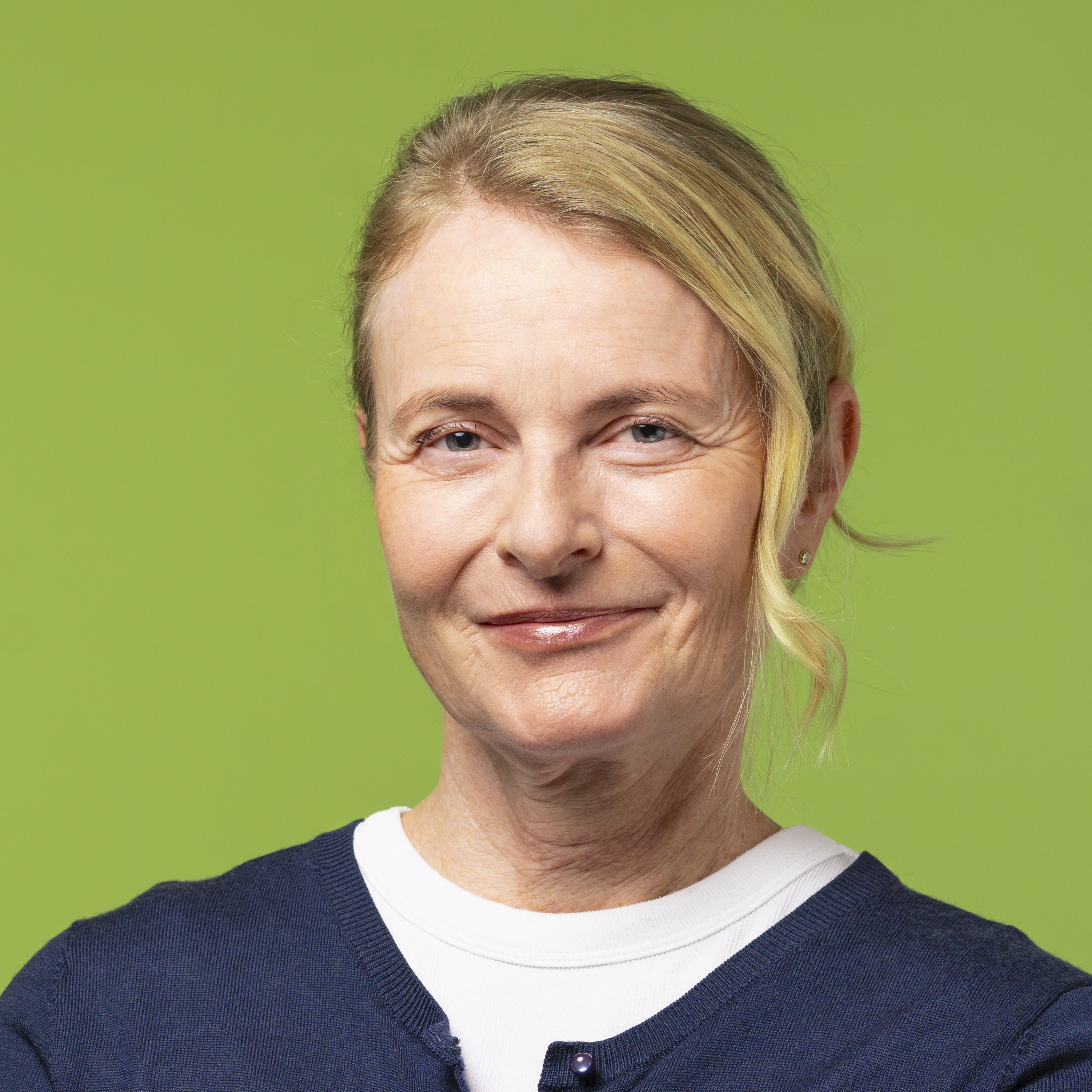 Frau mittleren Alters mit hellen, nach hinten gezogenen Haaren, die einen marineblauen Pullover über einem weißen Hemd trägt, steht vor einem einfarbig grünen Hintergrund, blickt in die Kamera und lächelt leicht.