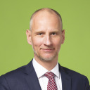 Ein Mann mit Glatze und gestutztem Bart, der einen dunklen Anzug, ein weißes Hemd und eine rot gestreifte Krawatte trägt, posiert vor einem schlichten grünen Hintergrund.