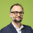 Ein Mann mit kurzen dunklen Haaren, einem Bart und einer Brille, der einen marineblauen Blazer und ein weißes Hemd trägt, steht vor einem einfarbig grünen Hintergrund.