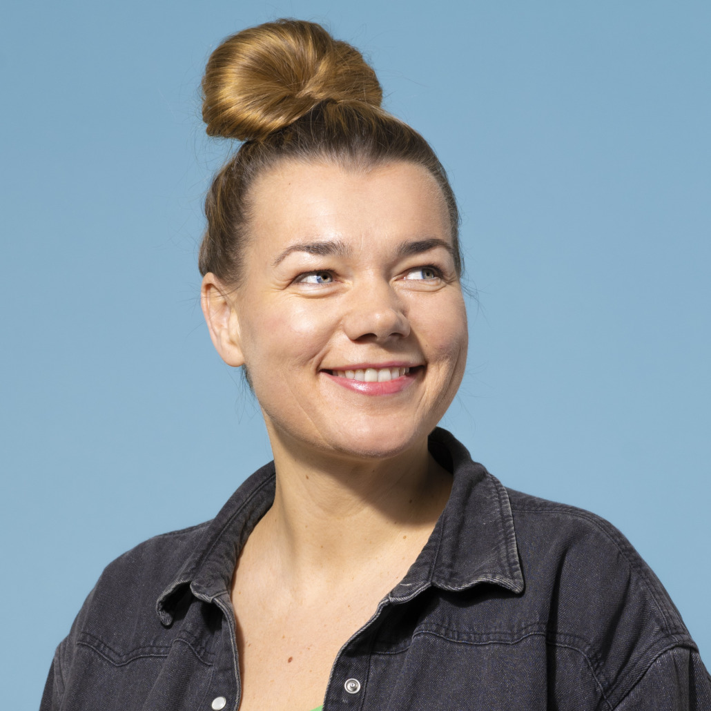 Portrait_ElisabethReinthaler Eine Frau mit heller Haut und braunem Haar in einem Dutt, die ein dunkles Jeanshemd trägt, lächelt und blickt zur Seite vor einem einfarbig blauen Hintergrund.