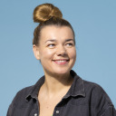 Eine Frau mit heller Haut und braunem Haar in einem Dutt, die ein dunkles Jeanshemd trägt, lächelt und blickt zur Seite vor einem einfarbig blauen Hintergrund.