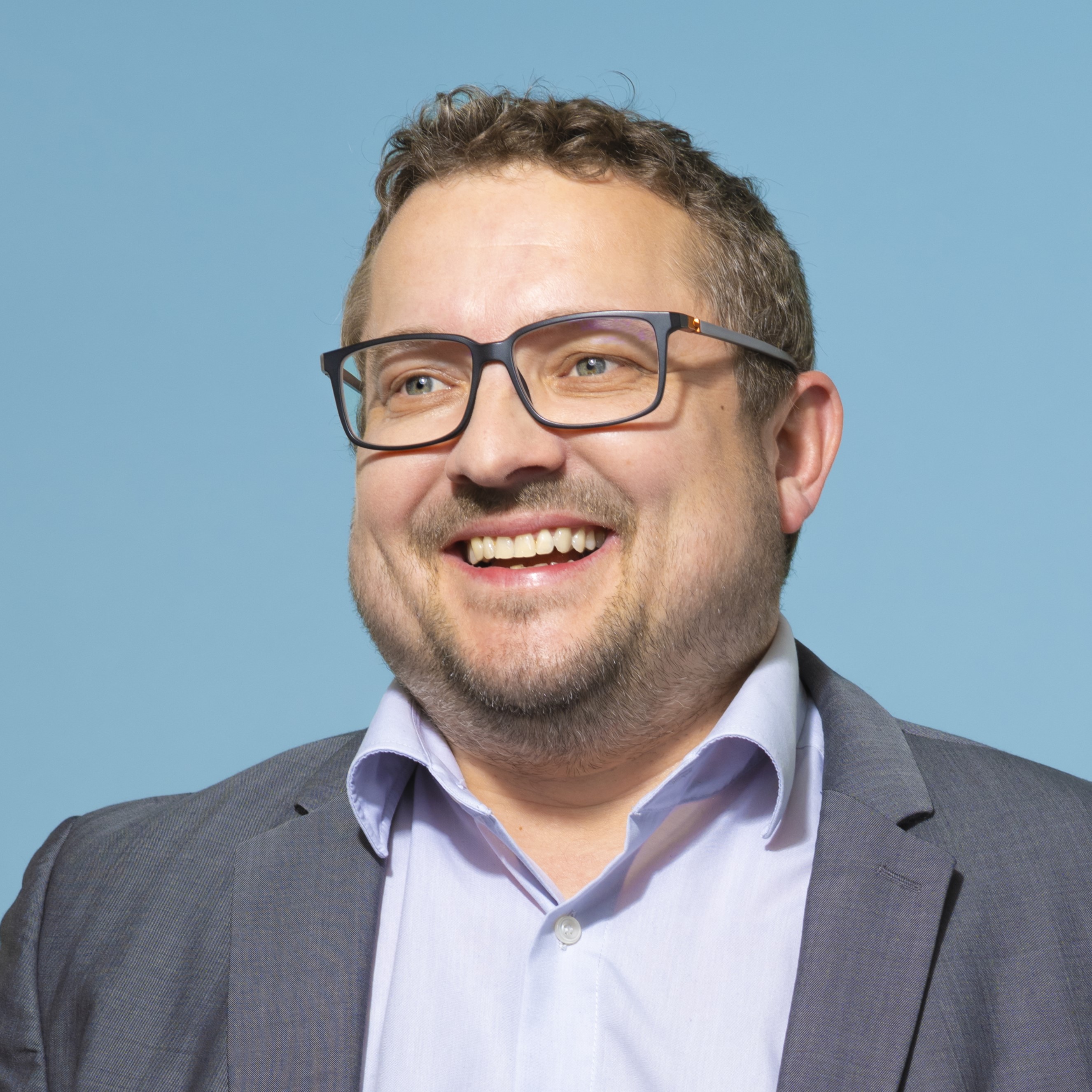 Ein Mann mit kurzen braunen Haaren und Brille, der eine Anzugsjacke und ein hellblaues Hemd trägt, lächelt vor einem einfarbig blauen Hintergrund.