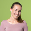 Eine Frau mit hellbraunem Haar in einem Pferdeschwanz, die ein hellrosa Oberteil trägt, lächelt vor einem einfarbig grünen Hintergrund.