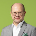 Ein Mann mittleren Alters mit Brille und Glatze trägt einen grauen Anzug, ein weißes Hemd und ein grünes Einstecktuch und steht vor einem schlichten grünen Hintergrund.