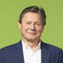 Ein älterer Mann mit kurzen braunen Haaren, der einen marineblauen Blazer und ein weißes Hemd trägt, lächelt vor einem schlichten hellgrünen Hintergrund.