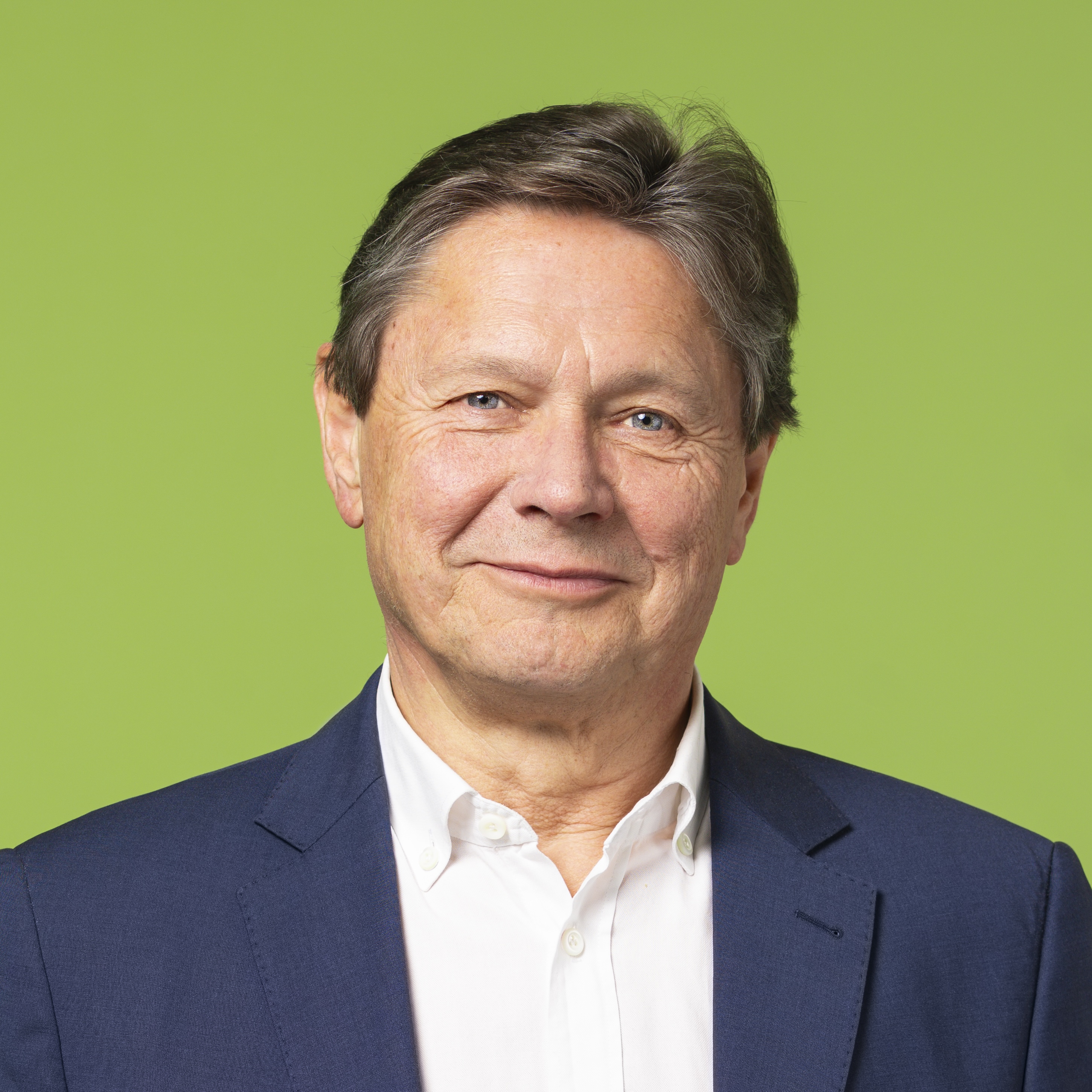 Ein älterer Mann mit kurzen braunen Haaren, der einen marineblauen Blazer und ein weißes Hemd trägt, lächelt vor einem schlichten hellgrünen Hintergrund.