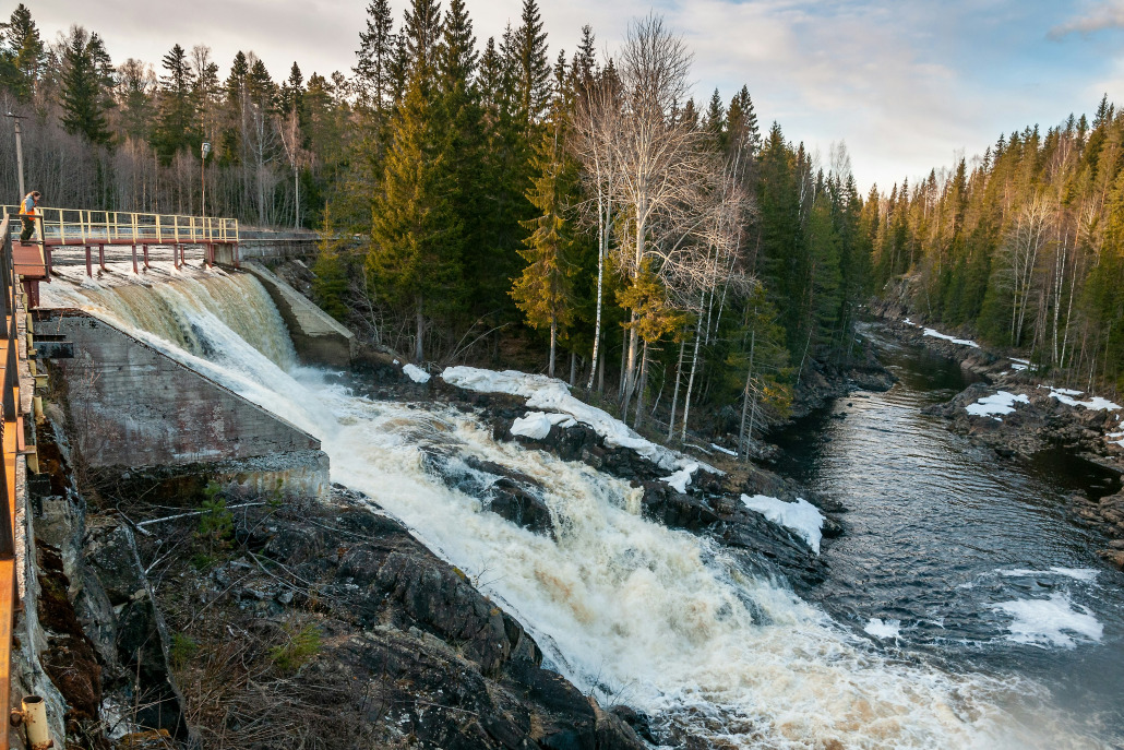 Ein kleiner Staudamm entlässt Wasser in einen felsigen Fluss, der von Nadelbäumen und Schneeflecken umgeben ist, in der Nähe eines Depotladens für emobilität LKW neu.