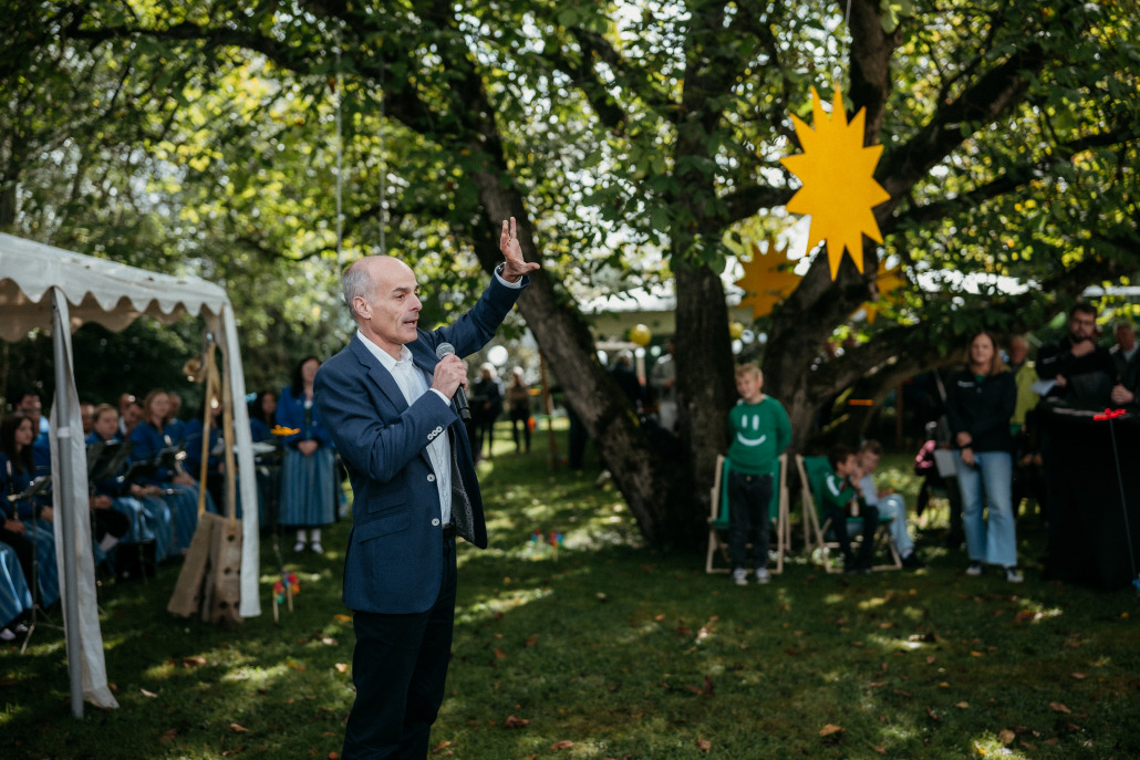 Beim Eröffnungsfest St. Veit spricht ein Mann im Anzug mit einem Mikrofon und gestikuliert zu einer Menschenmenge bei einer Veranstaltung im Freien unter Bäumen, die mit gelben Sternen geschmückt sind.