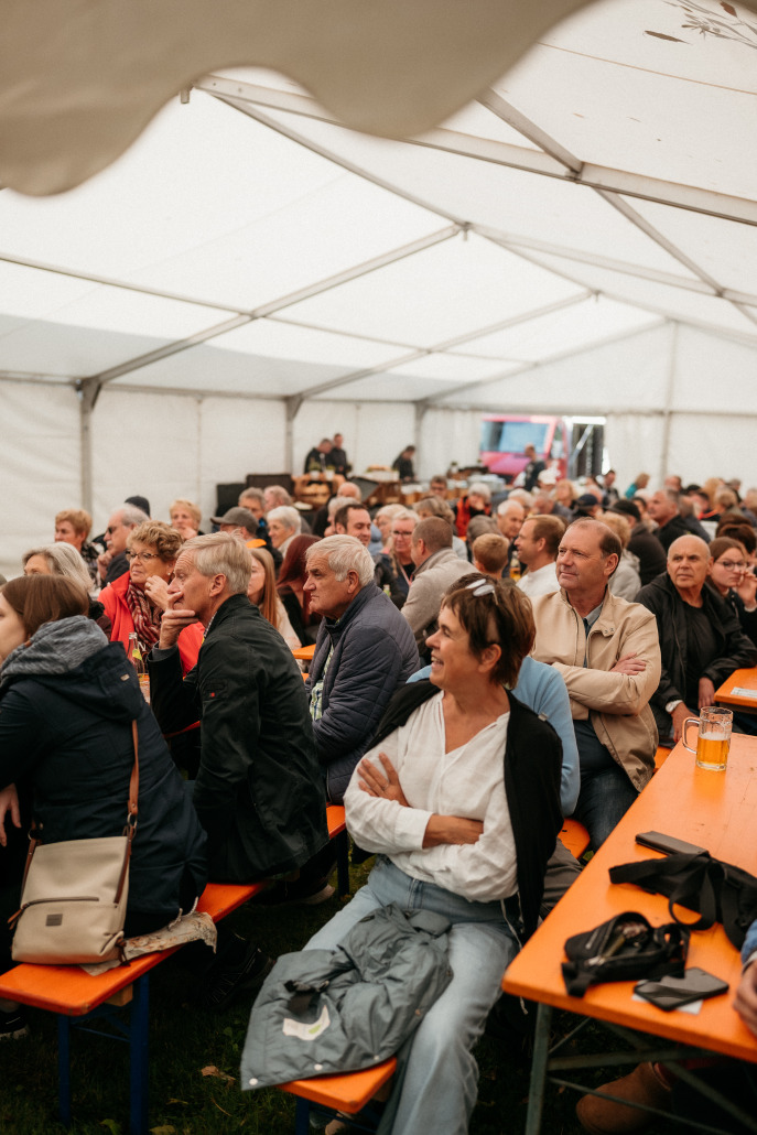 Eine Gruppe von Menschen sitzt auf orangefarbenen Bänken unter einem weißen Veranstaltungszelt auf dem Eröffnungsfest St. Veit und beobachtet aufmerksam etwas außerhalb des Bildes.